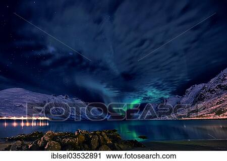 Stock Image - "Aurora over a fjord with snow-covered mountains and an illuminated village, Ersfjord, Tromso, â€ªTroms, Northern Norway, Norway, Europe". Fotosearch