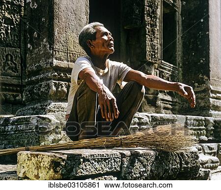 Stock Image - "Bayon temple, elderly man with broom squatting on the second terrace, Angkor Thom, Siem Reap Province, Cambodia, Asia". Fotosearch