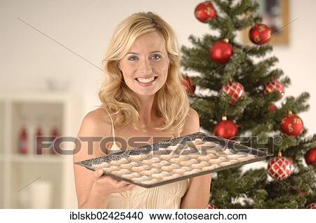 Stock Image - "Blonde woman, 20-30, wearing an evening dress while holding a baking tray with Christmas cookies in front of a Christmas tree". Fotosearch