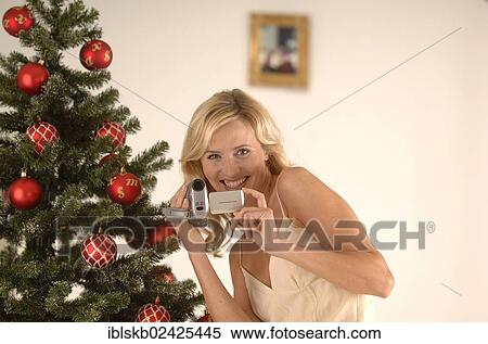 Stock Photography - "Blonde woman, 20-30, wearing an evening dress while filming with a camcorder in front of a Christmas tree". Fotosearch