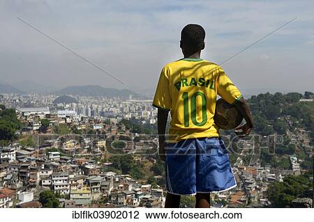 Stock Image - "Boy, 13 years, wearing the jersey of the Brazilian national team, holding a football, overlooking slums, favela, Rio de Janeiro, Brazil, South America". Fotosearch