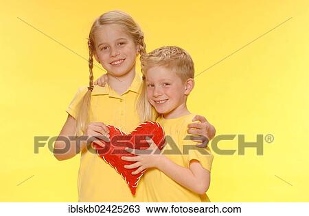 Stock Image - "Boy and girl, brother and sister, standing arm in arm, smiling, holding a red heart". Fotosearch