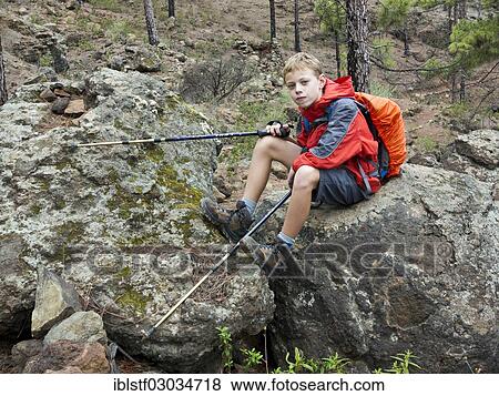 Stock Photo - "Boy holding hiking poles and wearing a backpack while sitting on a rock, Mogan, Gran Canaria, Spain, Europe". Fotosearch