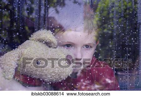 Picture - "Boy looking out of a window with water drops, holding a stuffed animal in his arms". Fotosearch