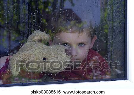 Stock Photograph - "Boy looking out of a window with water drops, holding a stuffed animal in his arms". Fotosearch