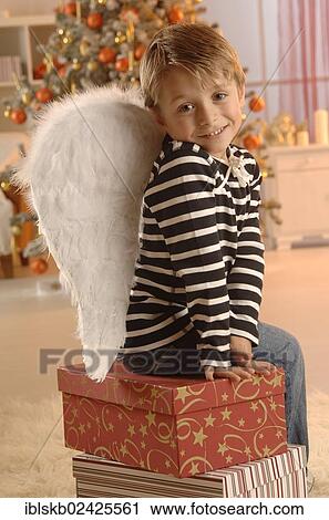 Stock Image - Boy wearing angel wings sitting on gifts in front of a Christmas tree. Fotosearch