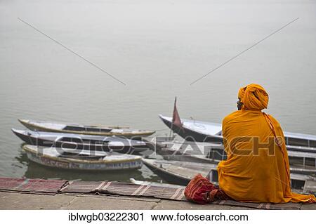 Stock Image - "Brahmin or Hindu priest sitting on a blanket on the banks of the Ganges River with several boats tied to the shore, Varanasi, Uttar Pradesh, India, Asia". Fotosearch