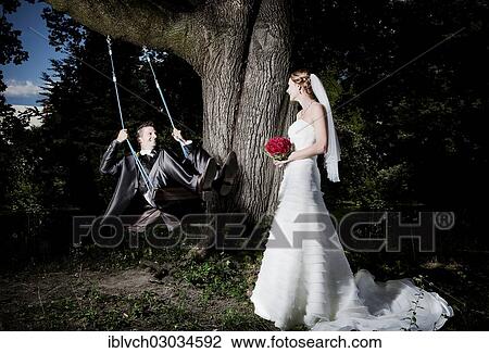 Stock Image - "Bride and groom, groom swinging on a swing, bride holding a bridal bouquet of red roses, Austria, Europe". Fotosearch