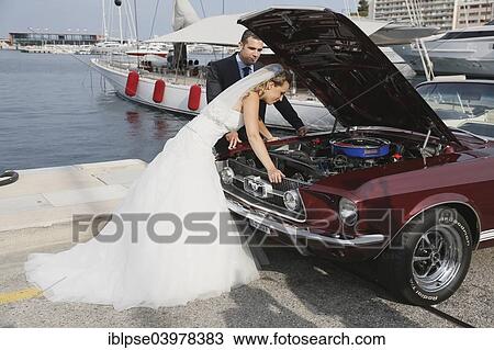 Stock Image - "Bride and groom looking into the engine compartment of a Ford Mustang after a breakdown, at a harbour". Fotosearch