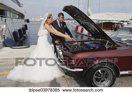 Stock Photography - "Bride and groom looking into the engine compartment of a Ford Mustang after a breakdown, at a harbour". Fotosearch