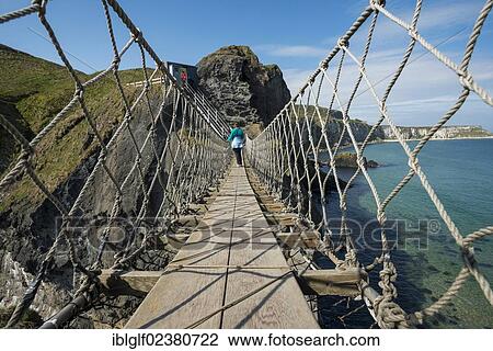 Stock Image - "Carrick-a-Rede Bridge, woman crossing the suspension bridge, Moyle, Northern Ireland, United Kingdom, Europe". Fotosearch
