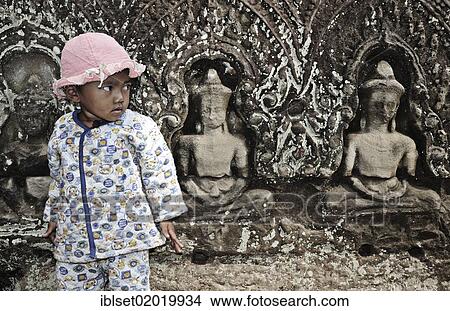 Picture - "Child standing in front of Buddha stone carvings, Angkor Wat, Siem Reap, Cambodia, Asia". Fotosearch