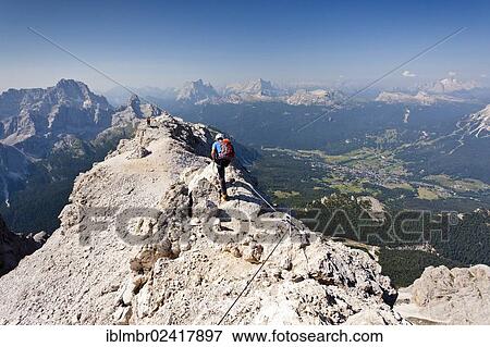 Stock Photo - Climber walking on the Marino Bianchi fixed rope route, on Monte Cristallo mountain, on the way to the summit of Cristallo di Mezzo mountain, Monte Pelmo mountain at the back, the village of Cortina below, Belluno, Dolomites, Italy, Europe. Fotosearch