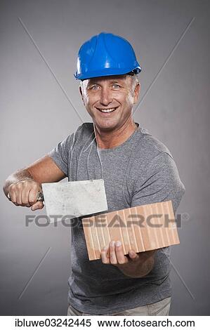 Stock Photography - "Construction worker wearing a hard hat, holding a trowel and a brick, Mannheim, Baden-Wurttemberg, Germany, Europe". Fotosearch