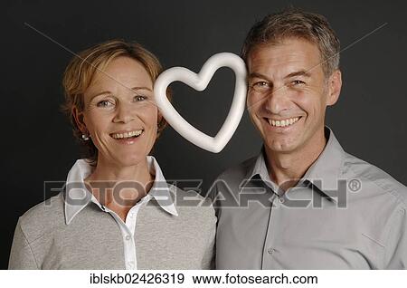 Stock Photo - "Couple, 35-45, standing next to each other, smiling and holding a styrofoam heart between them with their heads". Fotosearch