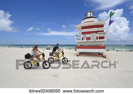 Stock Image - "Couple riding electric bicycles, Watchtower, The Jetty, Miami Rescue Tower, South Beach, Miami, Florida, USA". Fotosearch
