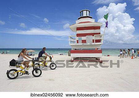 Stock Image - "Couple riding electric bicycles, Watchtower, The Jetty, Miami Rescue Tower, South Beach, Miami, USA". Fotosearch