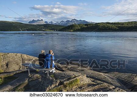 "Couple sitting on a bench looking out to the tidal Saltstraumen strait in the evening light, Borvasstindan mountain range at back, Saltstraumen, Skerstadfjord, Bodo, Nordland, Northern Norway, Norway, Europe" View Large Photo Image Stock Image - "Couple sitting on a bench looking out to the tidal Saltstraumen strait in the evening light, Borvasstindan mountain range at back, Saltstraumen, Skerstadfjord, Bodo, Nordland, Northern Norway, Norway, Europe". Fotosearch