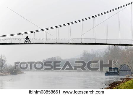 Stock Image - "Cyclist crossing a suspension bridge, water mill on the Weser river, Minden, North Rhine-Westphalia, Germany, Europe". Fotosearch