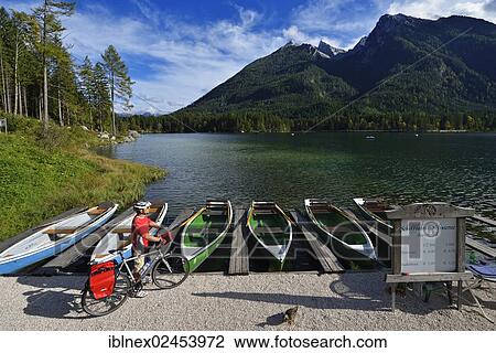 Stock Image - "Cyclist standing in front of boats at Hintersee lake, Ramsau, Berchtesgaden, Upper Bavaria, Bavaria, Germany, Europe". Fotosearch