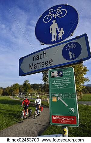 Stock Image - "Cyclists riding electric bicycles below a signpost with bicycle routes, Fraham, Lake Grabensee, Salzburg Lake District, Salzburg, Austria, Europe". Fotosearch