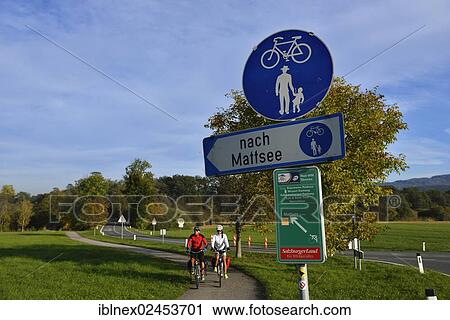 Stock Image - "Cyclists riding electric bicycles below a signpost with bicycle routes, Fraham, Lake Grabensee, Salzburg Lake District, Salzburg, Austria, Europe". Fotosearch