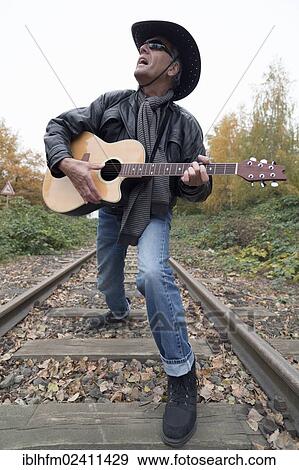 "Elderly man wearing jeans and a black leather jacket with a black cowboy hat and a western guitar singing on a train track, Hagen, Lower Saxony, Germany, Europe" View Large Photo Image Stock Photo - "Elderly man wearing jeans and a black leather jacket with a black cowboy hat and a western guitar singing on a train track, Hagen, Lower Saxony, Germany, Europe". Fotosearch