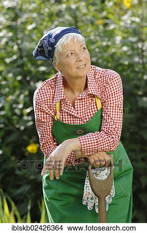 Picture - "Elderly woman, gray-haired, 55-65, wearing gardening clothes and leaning against a spade in a garden". Fotosearch