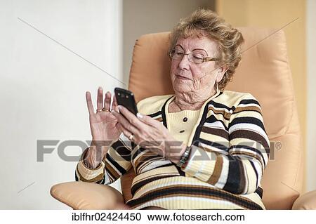 Stock Image - Elderly woman sitting in an armchair and using a smartphone, Germany, Europe. Fotosearch
