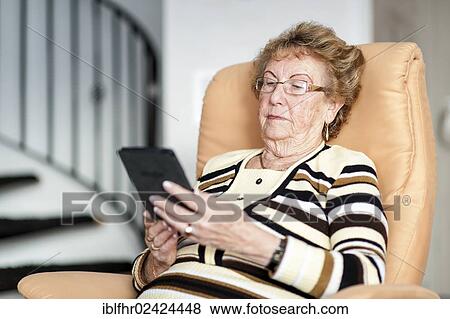 Stock Photo - Elderly woman sitting in an armchair and reading an eBook, Germany, Europe. Fotosearch
