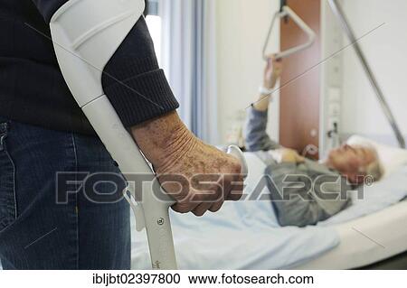 Stock Image - Elderly woman standing on a crutch in front of an elderly man lying in a hospital bed. Fotosearch