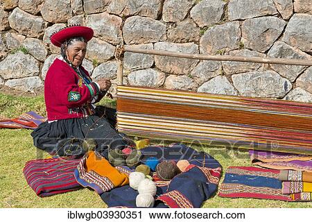 Stock Image - "Elderly woman wearing a hat, Quechua Indian in traditional dress sitting on the floor working on the stretcher of a loom, Cinchero, Urubamba Valley, Peru, South America". Fotosearch