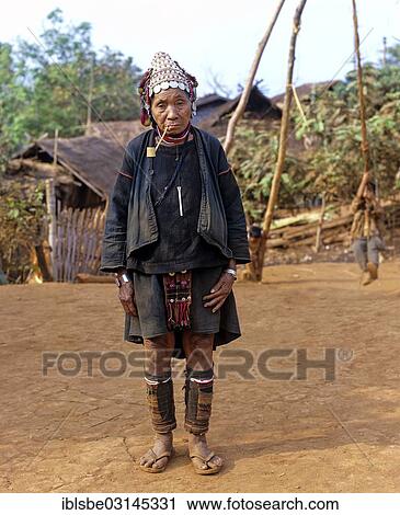 Stock Image - "Elderly woman with a whistle standing in front of bamboo huts in an Akha village, Chiang Rai Province, Northern Thailand, Thailand, Asia". Fotosearch