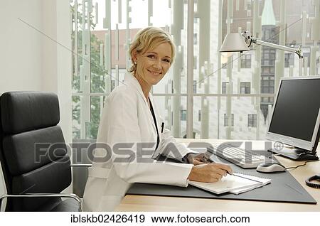 Stock Photo - Female doctor in white coat sitting at her desk. Fotosearch