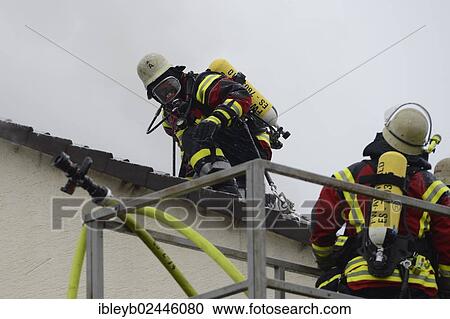 Stock Image - "Firefighters wearing breathing equipment while extinguishing a roof fire, removing the roof tiles to access the roof, Aichwald, Baden-Wurttemberg, Germany, Europe". Fotosearch