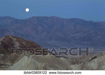 Stock Image - "Full moon over the Panamint Range and the Death Valley at dawn, seen from Zabriskie Point, the badlands of Gower Gulch in the foreground, Death Valley National Park, California, USA". Fotosearch