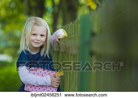 Stock Image - "Girl, 4 years old, holding a suitcase leaning against a wooden fence". Fotosearch