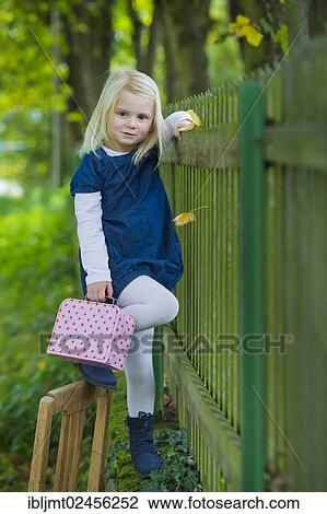 Stock Image - "Girl, 4 years old, holding a suitcase leaning against a wooden fence". Fotosearch