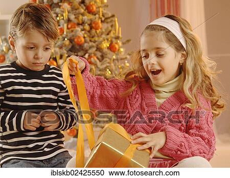 Stock Image - Girl and boy unpacking a gift in front of a Christmas tree. Fotosearch