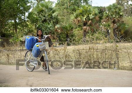 Picture - "Girl transporting two canisters with water from the reservoir of the nearby pagoda on her bike to her home, Trapang Village, Bathi District, Takeo Province, Cambodia, Asia". Fotosearch