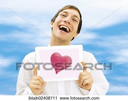 Stock Image - "Happy man holding an iPad with a heart, against a blue sky, romantic, in love". Fotosearch