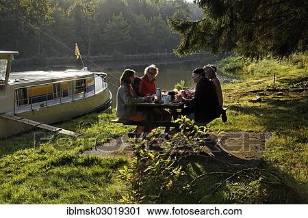 Stock Image - "Holidaymakers having breakfast on a picnic table, morning mood, houseboat on the Canal des Vosges, formerly Canal de l'Est, landing in the forest at PK 115, Thunimont, Epinal, Lorraine, Departement Vosges, France, Europe". Fotosearch
