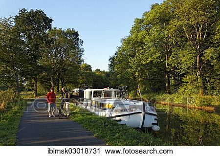 Stock Image - "Houseboat on the Canal des Vosges, formerly the Canal de l'Est, Golbey flight of locks, cyclist on the towpath, Les Forges, Epinal, Lorraine, Departement Vosges, France, Europe". Fotosearch