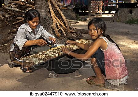 Stock Image - "Khmer woman and a girl filling caramelized palm sugar into small moulds made of strips of palm leaves for the production of traditional palm sugar candies, Siem Reap, Cambodia, Southeast Asia, Asia". Fotosearch