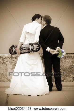 "Kissing wedding couple seen from behind, the bride is holding a skateboard, the groom is wearing handcuffs and holding a bridal bouquet" View Large Photo Image Stock Image - "Kissing wedding couple seen from behind, the bride is holding a skateboard, the groom is wearing handcuffs and holding a bridal bouquet". Fotosearch