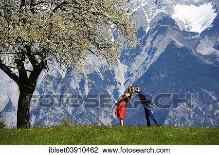 Stock Image - "Lovers kissing next to a flowering tree, in the spring, mountains at the back, Tyrol, Austria, Europe". Fotosearch