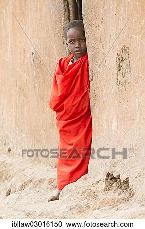 Stock Image - "Maasai child wearing traditional dress looking out of a mud hut, Massai Mara, Distrikt Narok, Serengeti, Rift Valley province, Kenya, Africa". Fotosearch