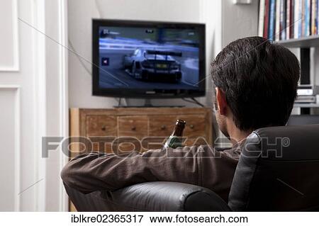 Man holding a bottle of beer while sitting on a leather sofa in front of a TV watching motorsport View Large Photo Image Stock Photo - Man holding a bottle of beer while sitting on a leather sofa in front of a TV watching motorsport. Fotosearch