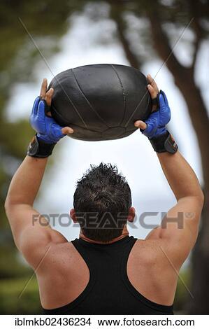 Picture - Man holding a medicine ball over his head while exercising in a park. Fotosearch