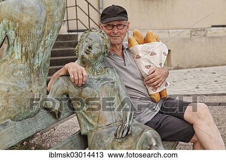 Stock Image - "Man holding baguettes sitting beside a sculpture in front of the Town Hall, Port-sur-Saone, Vesoul, Region Franche-Comte, Dep. Haute-Saone, France, Europe". Fotosearch